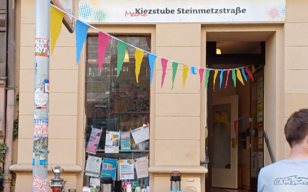 Eingang der „Kiezstube Steinmetzstraße“ mit bunter Wimpelkette; vor dem Schaufenster stehen Thermoskannen und Infomaterial, im Fenster hängen Flyer.