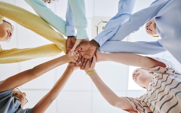 Fünf Personen stehen im Kreis und legen ihre Hände übereinander in die Mitte. Die Hände sind unterschiedlich und symbolisieren Gemeinschaft und Zusammenhalt. Im Hintergrund sind lächelnde Gesichter zu erkennen.