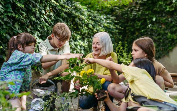 Eine Frau mit weißem Haar und gelbem Oberteil sitzt lachend im Freien und zeigt gemeinsam mit vier Kindern auf eine blühende Pflanze mit gelben Blüten. Im Hintergrund eine dichte grüne Hecke; eines der Kinder hält eine Gießkanne.