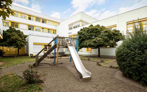 Innenhof‑Spielplatz vor weißem Gebäude mit gelben Fenstern: Kletterturm mit Rutsche, Sitzbänke und Bäume rundherum.