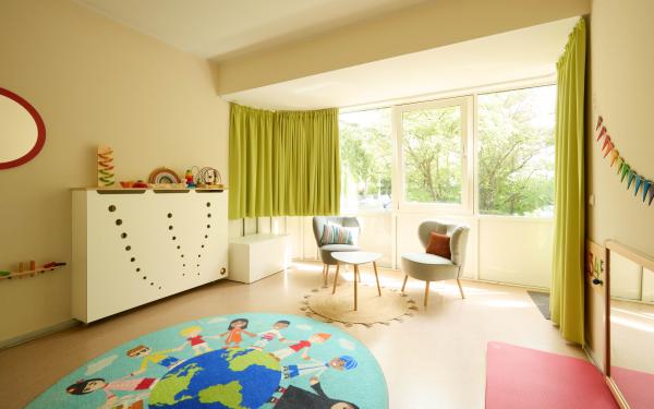 Heller Spiel- und Aufenthaltsraum mit grünen Vorhängen, niedrigen Regalen mit Spielmaterial und einem bunten Teppich. Rechts stehen kleine Tische und Stühle.