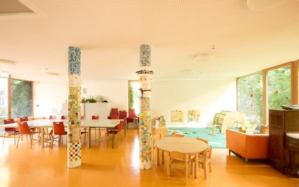 Großer Raum für Familienangebote mit hellem Boden und zwei bunt gekachelten Säulen. Links Tische und rote Stühle, rechts Spielbereich mit grünem Teppich, Kletterbogen und Wandspieltafeln; große Fenster ermöglichen Blick ins Grüne.