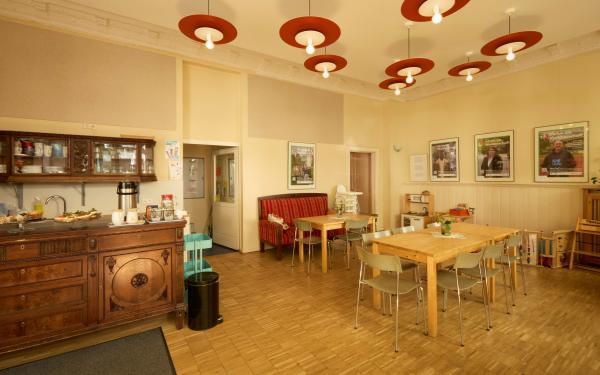 Gemütliches Café mit Holzboden: links eine antike Theke mit Geschirr und Kaffeemaschine, in der Mitte Holztische mit Stühlen und eine rote Polsterbank, an der Decke runde Pendelleuchten; an der Wand hängen Poster, rechts stehen Kinder­spielzeuge und Hochstühle.