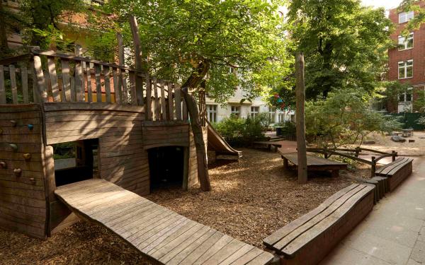 Schattiger Kita‑Hof mit großen Spielplatz aus Holz, Sitzbänken, Bäumen und Sträuchern.