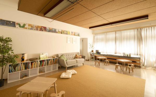 Heller Leseraum mit großem Fenster, beiger Teppichfläche und niedrigen Regalen voller Bilderbücher; vorne kleine Holztische, hinten ein graues Sofa und Sitzbank am Fenster.