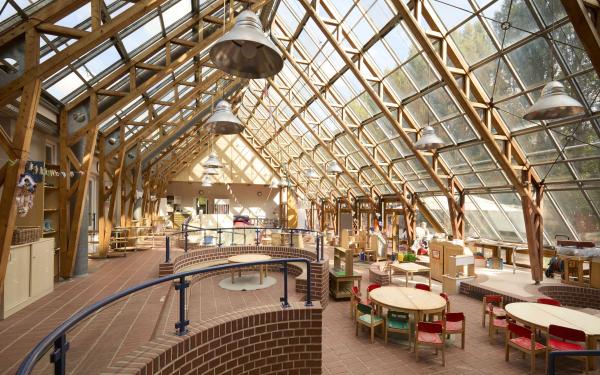 Großes lichtdurchflutetes Glashaus: Tische mit kleinen Stühlen, verschiedene Spiel‑ und Werkbereiche, große Hängelampen unter der Glasdecke.