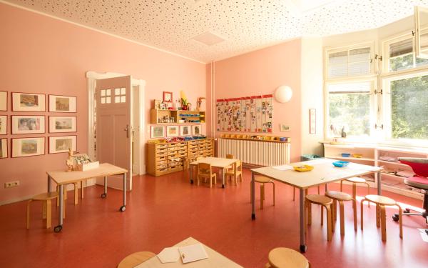Ein Atelier mit rosafarbenen Wänden ist mit niedrigen Tischen und Stühlen ausgestattet. Gerahmte Bilder und Kinderarbeiten schmücken die Wände, und in Regalen sind Materialien und Spielsachen verstaut.
