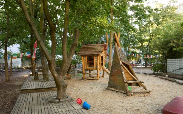 Schattiger Kita‑Spielplatz mit vielen Bäumen: Sandfläche mit Holzhäuschen und Spielzeugen; im Hintergrund bunte Wimpelkette und ein Fußballtor.