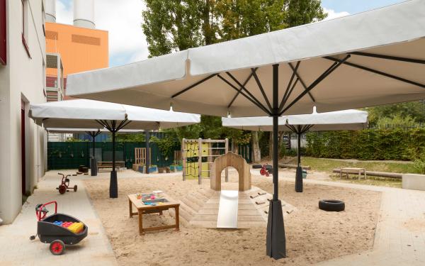 Heller Sandspielbereich mit großen Sonnenschirmen, kleinem Kletter‑ und Rutschhügel, Werkbanktisch und Dreirädern; Hochbeete und Sitzbänke am Rand.
