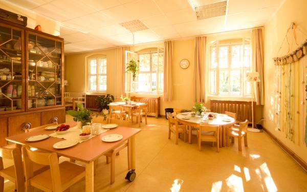 Ein heller Essbereich ist mit langen Holztischen und Stühlen eingerichtet; auf den Tischen steht Geschirr und große Fenster lassen Tageslicht herein.