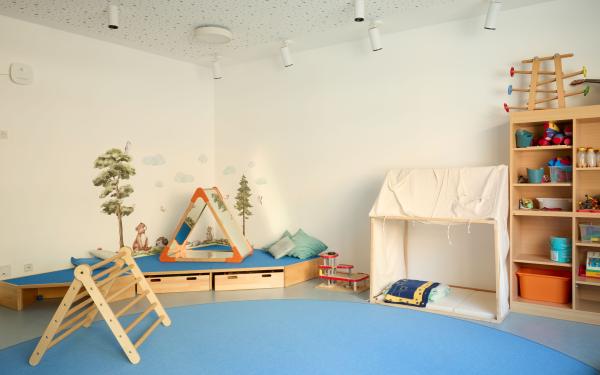 Heller Spielraum im Nestbereich mit großem blauem Teppichkreis, hinten eine niedrige gepolsterte Ecke mit Wandmalerei und Spielzeug; rechts ein kleines Stoffzelt mit Matten und Decken sowie ein offenes Regal mit sortierten Spielmaterialien.