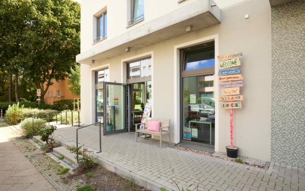Eingang eines modernen Nachbarschaftszentrums mit beiger Fassade, Glastür, Holzbank mit rosa Kissen und mehrsprachigem Willkommensschild an der Wand. Gepflasterte Terrasse mit Grünflächen und Bäumen im Hintergrund.