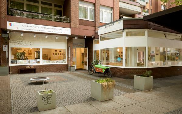  Außenansicht des Nachbarschaftszentrums in einem roten Klinkerbau. Große Schaufenster geben den Blick in helle Räume frei; vor dem Eckfenster steht ein grünes Lastenrad. Davor ein kleiner gepflasterter Platz mit Pflanzkübeln.